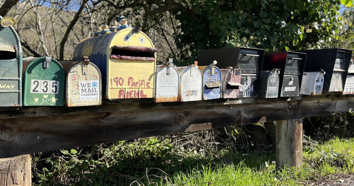 A row of colorful, weathered rural mailboxes lined up on a long wooden rail in the sun
