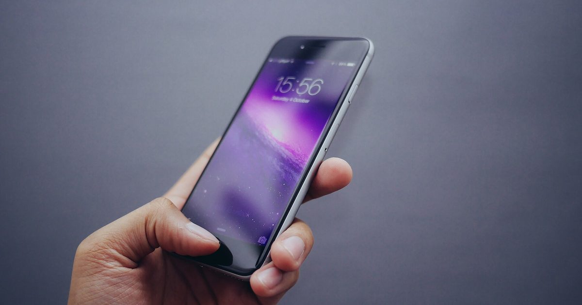 Hand holding a smartphone with a purple lock screen showing the time 15:56, against a plain grey background