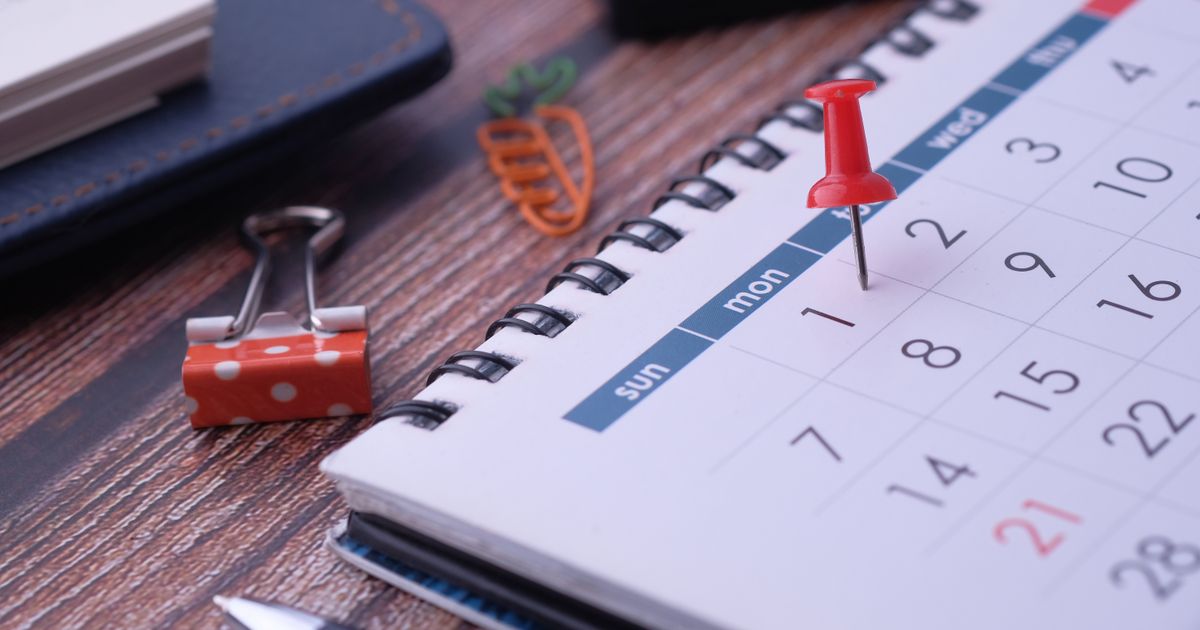 Red push pin marking a date on a desk calendar, next to a binder clip and paper clips on a wooden desk