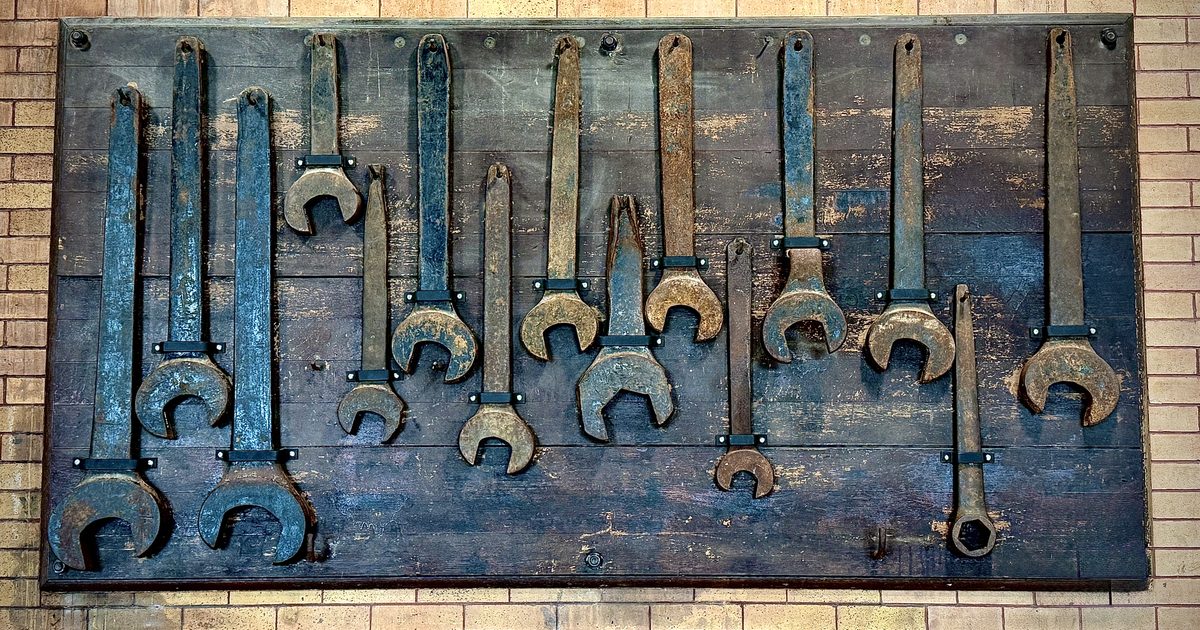 Antique metal wrenches of different sizes hanging in two uneven rows on a dark wooden board against a brick wall