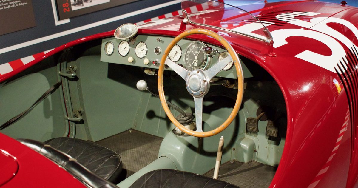 The cockpit of a vintage red racing car with a wooden steering wheel and a row of round dashboard gauges