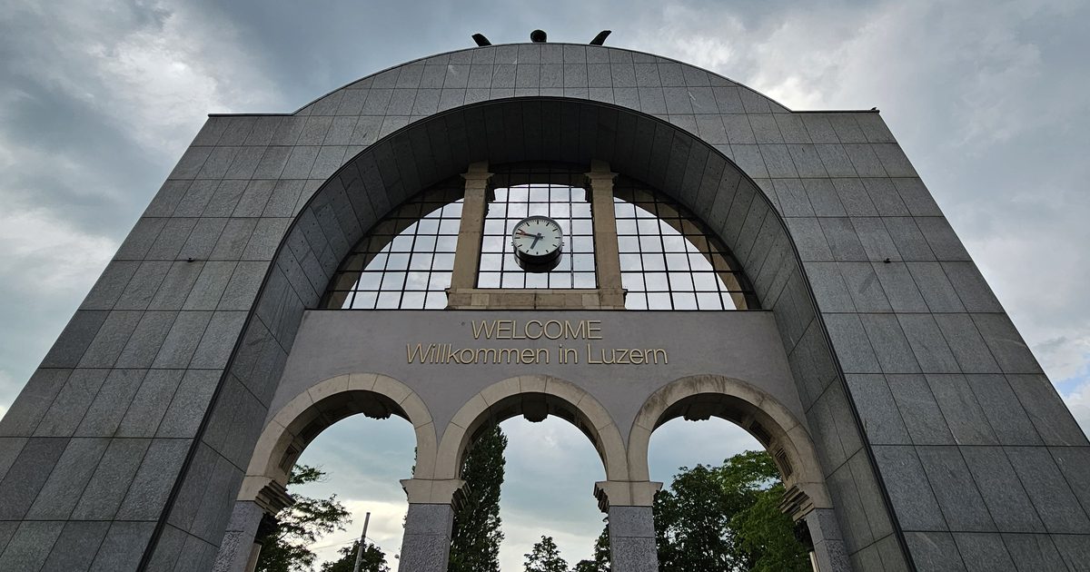 A stone arch with the words Welcome and Willkommen in Luzern in gold letters under a station clock and a cloudy sky