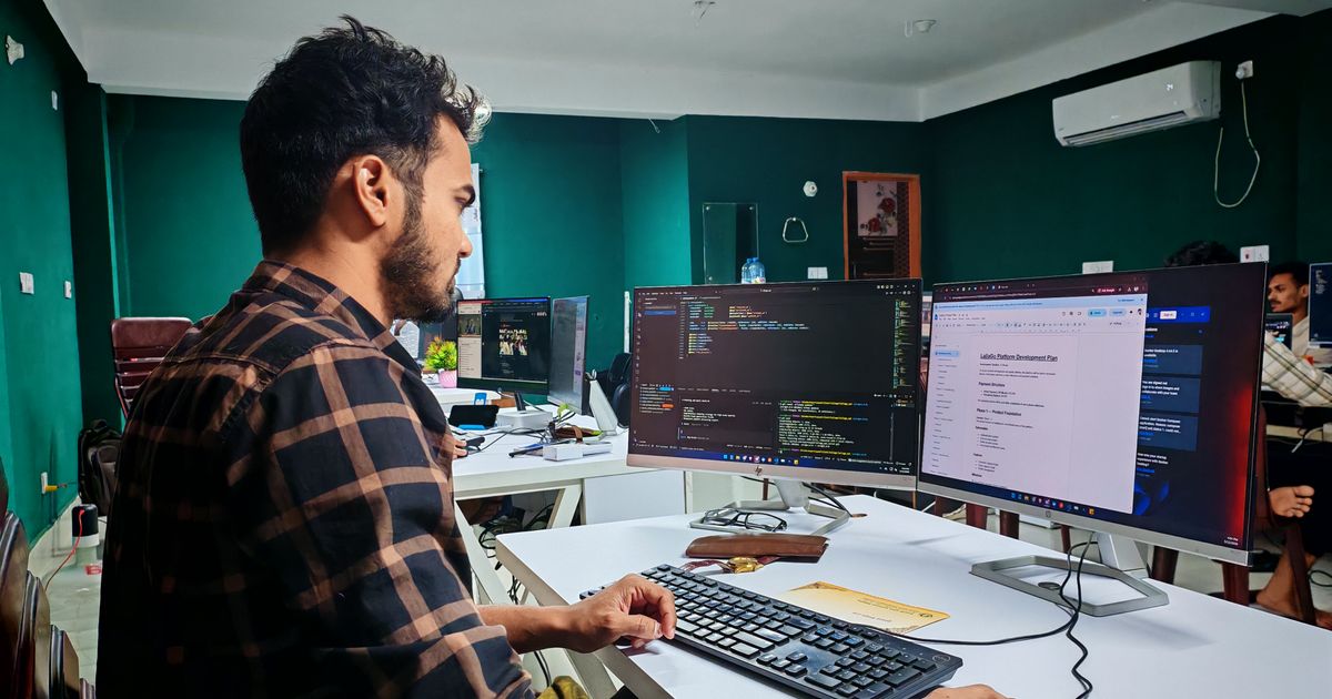Developer at a desk in an open office working across two monitors, one showing a code editor and the other a project plan