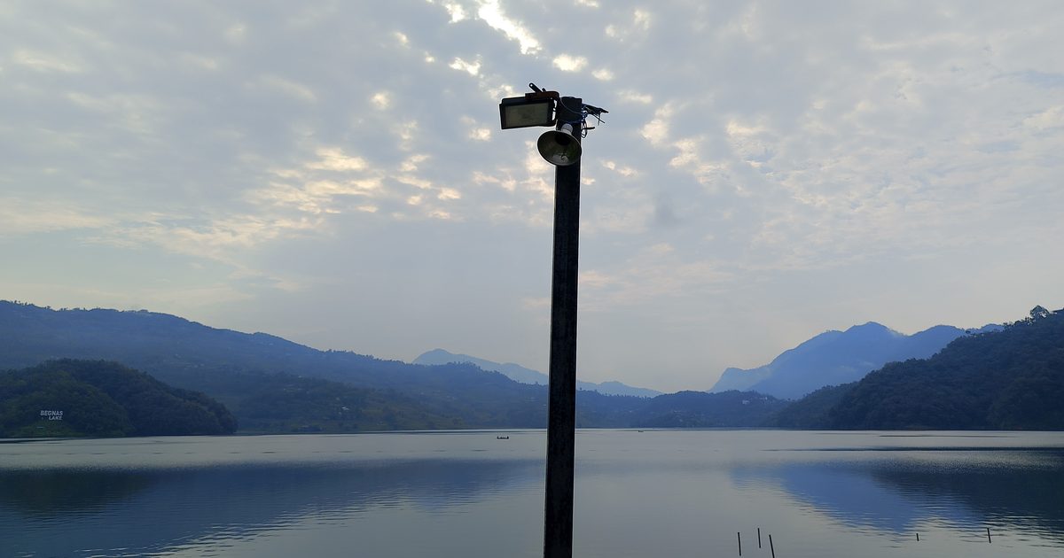A lamp post with a loudspeaker standing in front of a calm lake ringed by misty blue hills under a cloudy sky