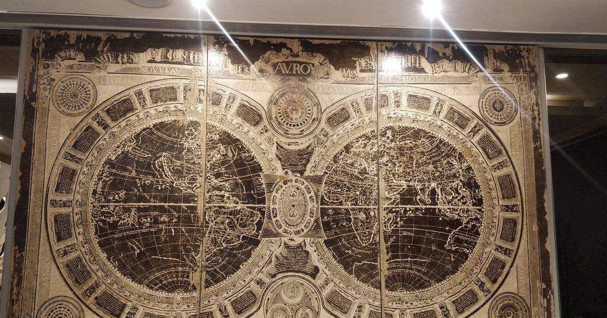 A large antique double-hemisphere world map in gold and black mounted on a wall under bright ceiling lights
