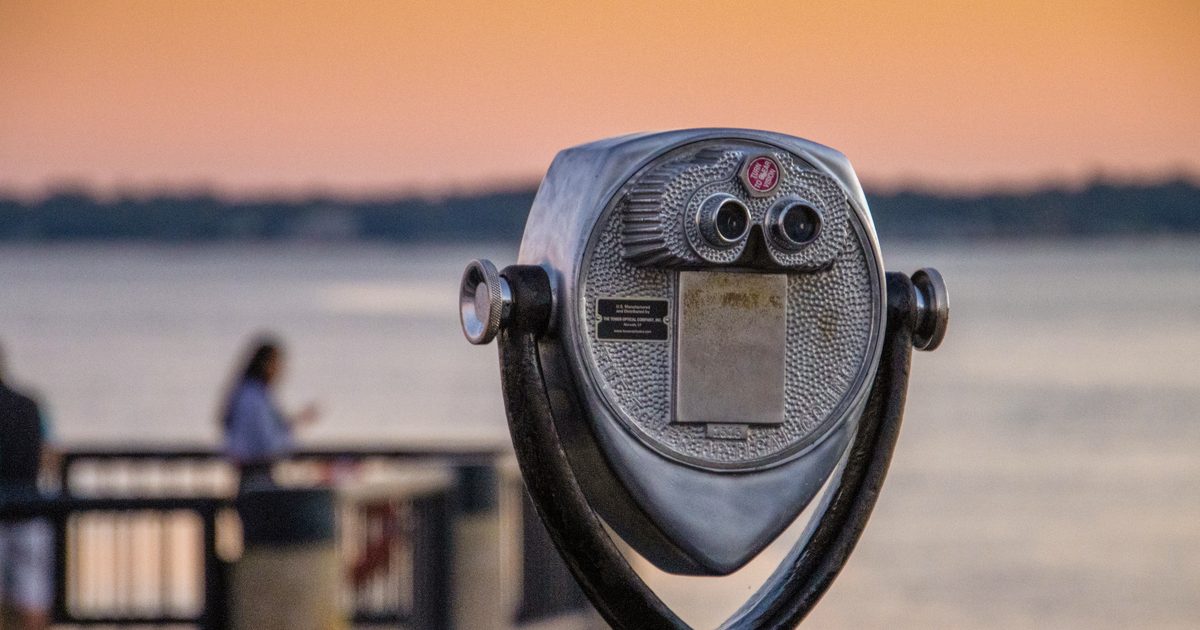 Close-up of a coin-operated sightseeing binocular viewer on a waterfront promenade at sunset, with blurred people and water behind it