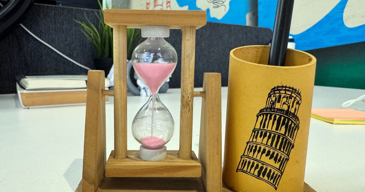 Wooden hourglass with pink sand running into the lower bulb, standing next to a yellow pen holder on an office desk