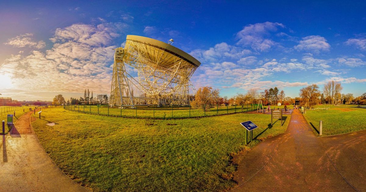 The white dish of the Lovell radio telescope at Jodrell Bank under a blue sky, with grass and footpaths in the foreground