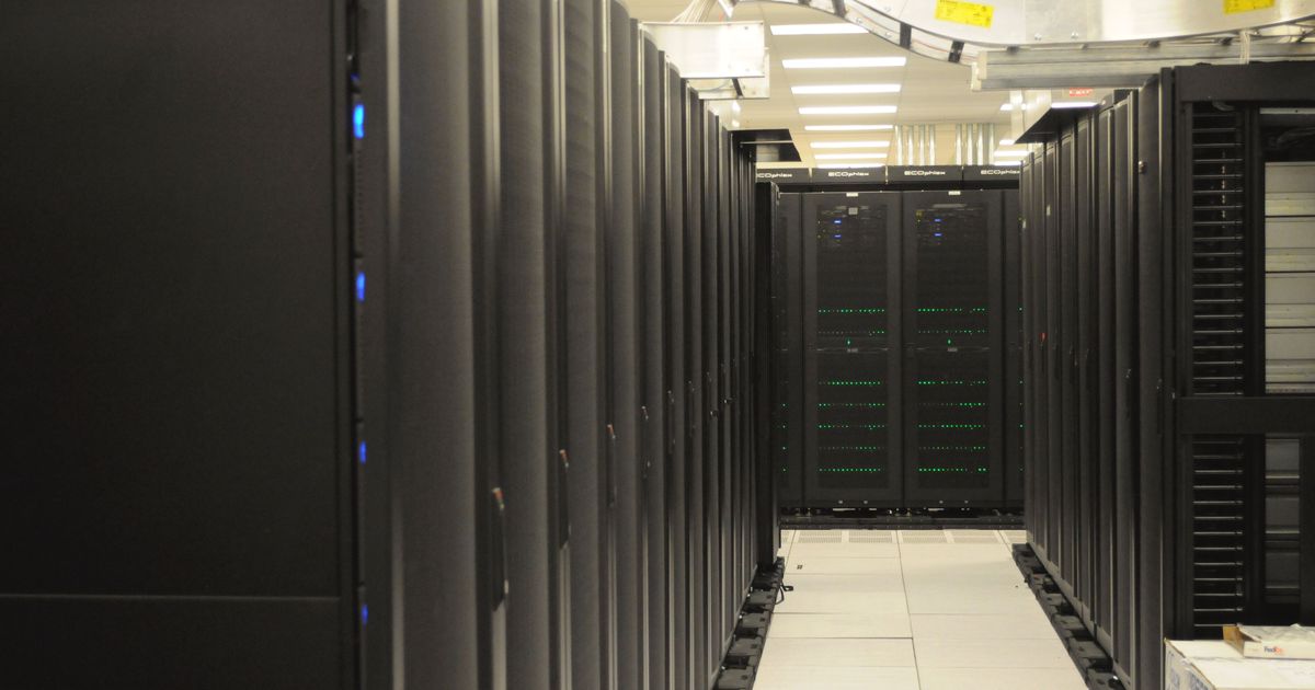 Long aisle between rows of black server racks in a data center, with green status lights and an overhead cable tray
