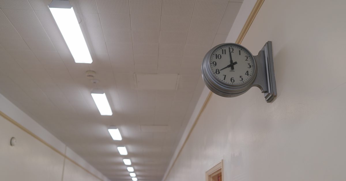 A round silver wall clock mounted on a pale hallway wall, with a long row of ceiling lights fading into the distance