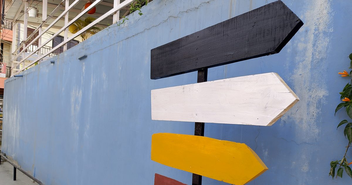 A wooden signpost on a pale blue wall with stacked arrows painted black, white and yellow, all pointing to the right