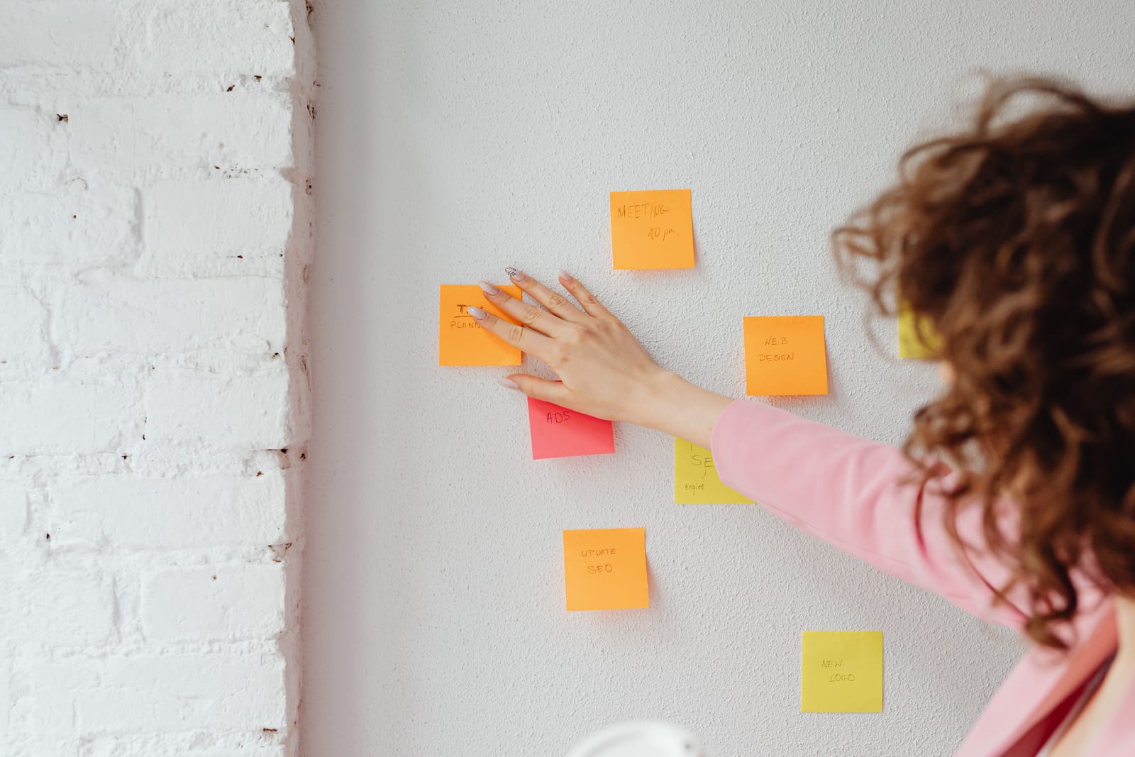 Woman in a pink shirt sticking handwritten orange, pink and yellow sticky notes onto a white wall
