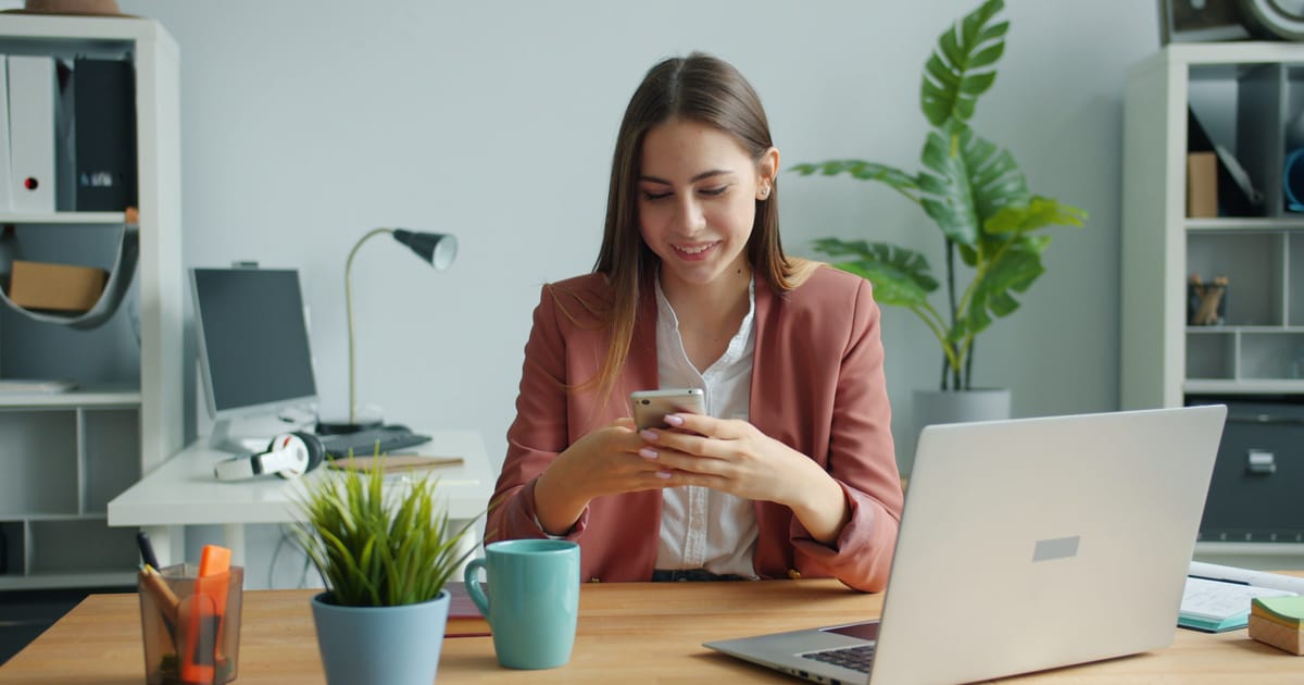 Smiling woman reading a notification on her phone at an office desk, with an open laptop and a coffee mug in front of her