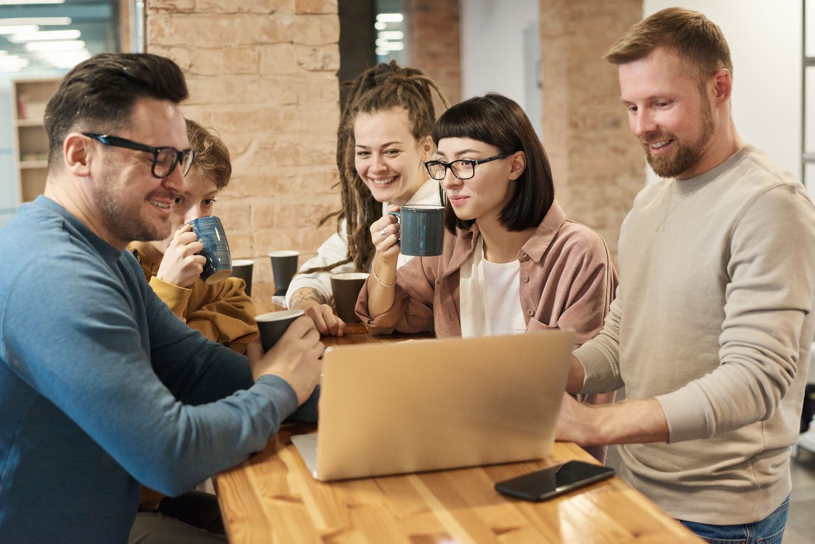 Five colleagues with coffee mugs gathered around one open laptop at a wooden table