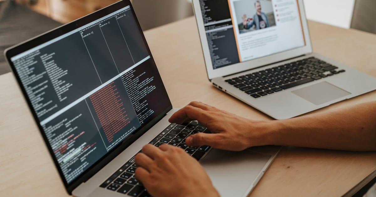 Developer typing on a laptop with terminal panes and build output open, a second laptop beside it on a wooden desk